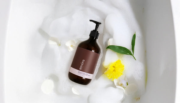 Suvéra Hand & Body Wash Patchouli & Amber Vanilla bottle floating in a foam-filled bathtub, overhead view with soft bubbles and warm lighting, evoking spa-like relaxation and natural skincare.