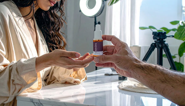 Male hand offering a skincare serum to a woman in a robe getting ready at her vanity; blurred tripod with ring light and phone filming in the background; influencer vibe.