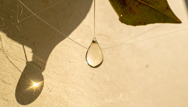 Water droplet and small leaf on a thin acrylic ledge; warm side light casting soft shadows on a matte limestone wall.
