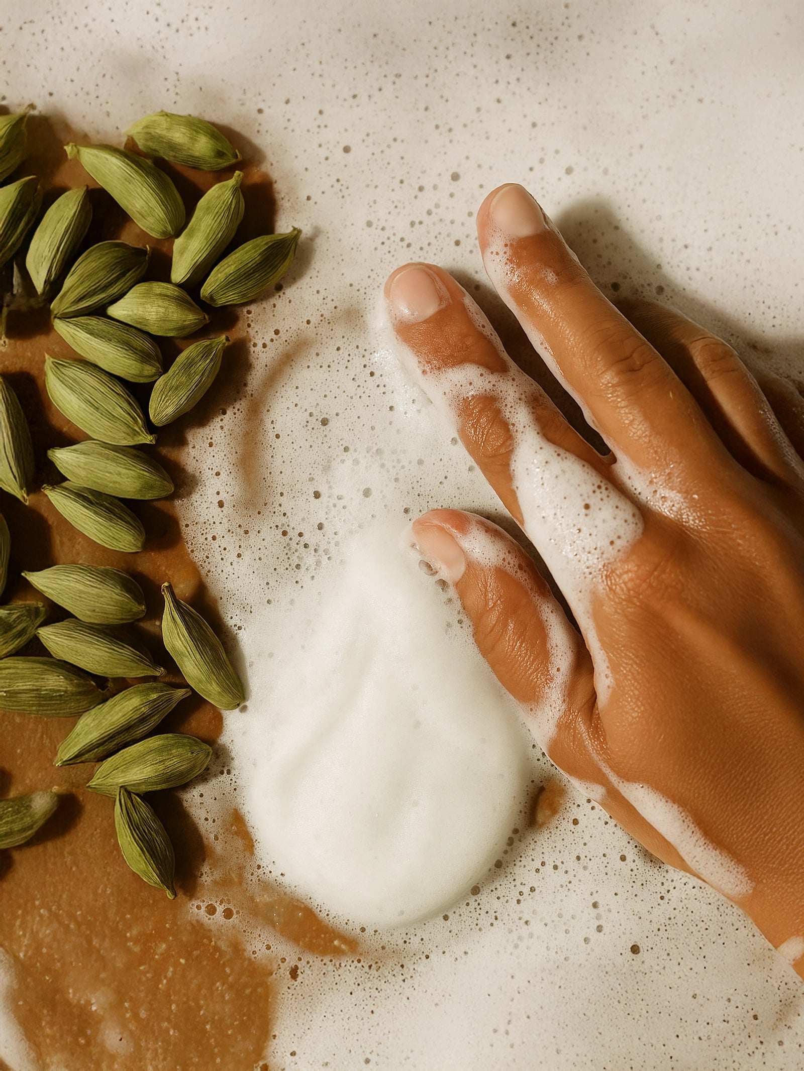 Hand with soap foam next to cardamom pods on a textured surface Suvéra hand and body wash with creamy white foam and natural cardamom tones, gentle cleansing skincare, sustainable self care beauty