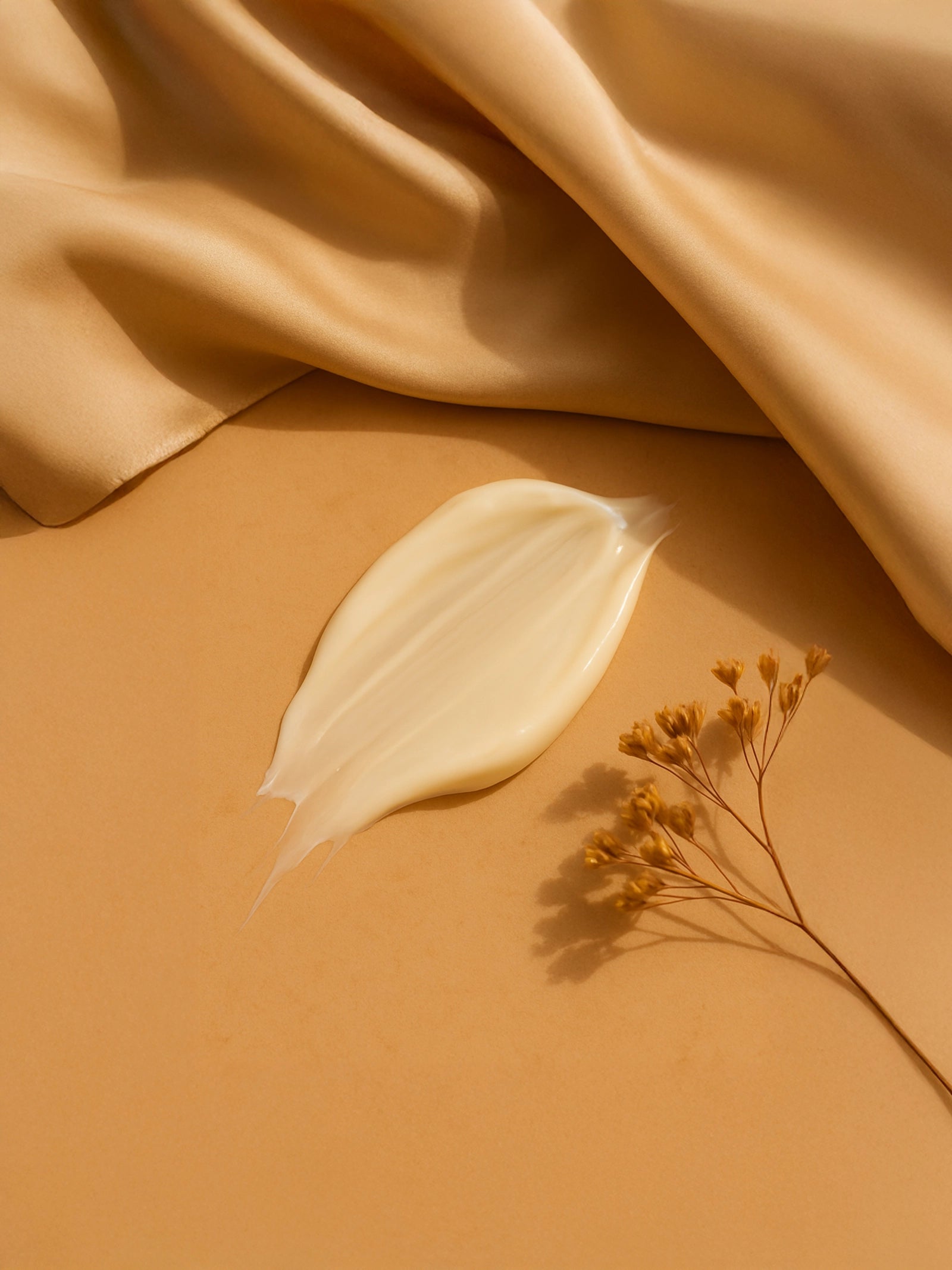 Creamy white droplet on a beige fabric background with a small yellow flower. Close-up of skincare serum drops beside champagne silk fabric and dried flowers on a neutral backdrop