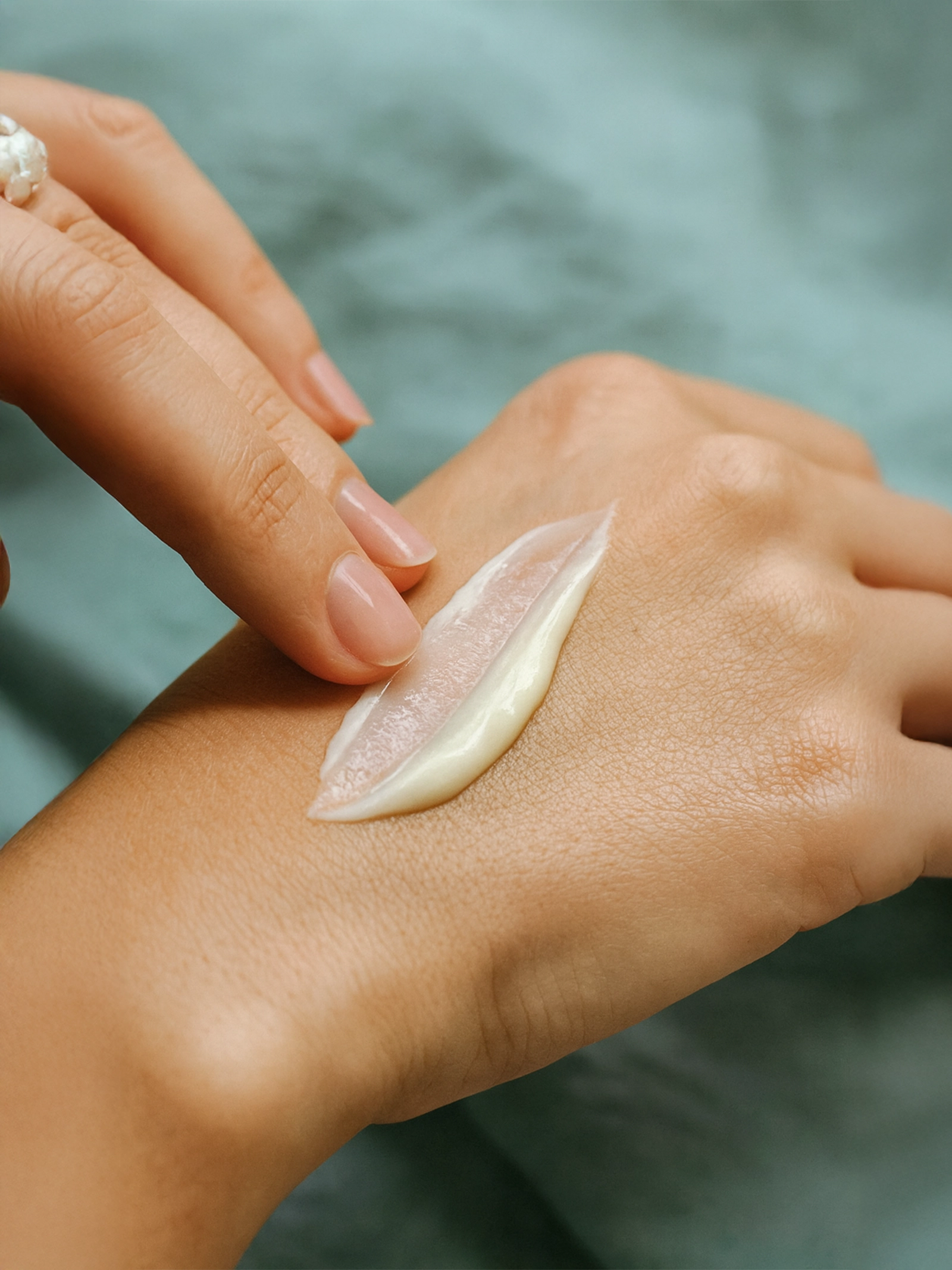 Hand applying a small amount of cream to another hand against a blurred background Close-up of fingertips spreading a pearlescent cream across the back of a hand over soft teal fabric.