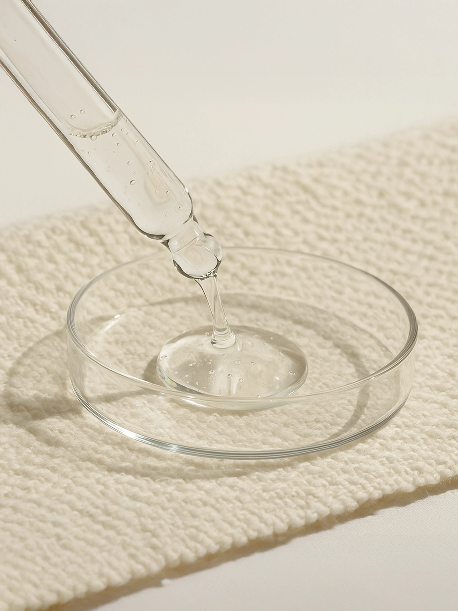Glass dropper with clear liquid being poured into a round glass dish on a textured beige surface.