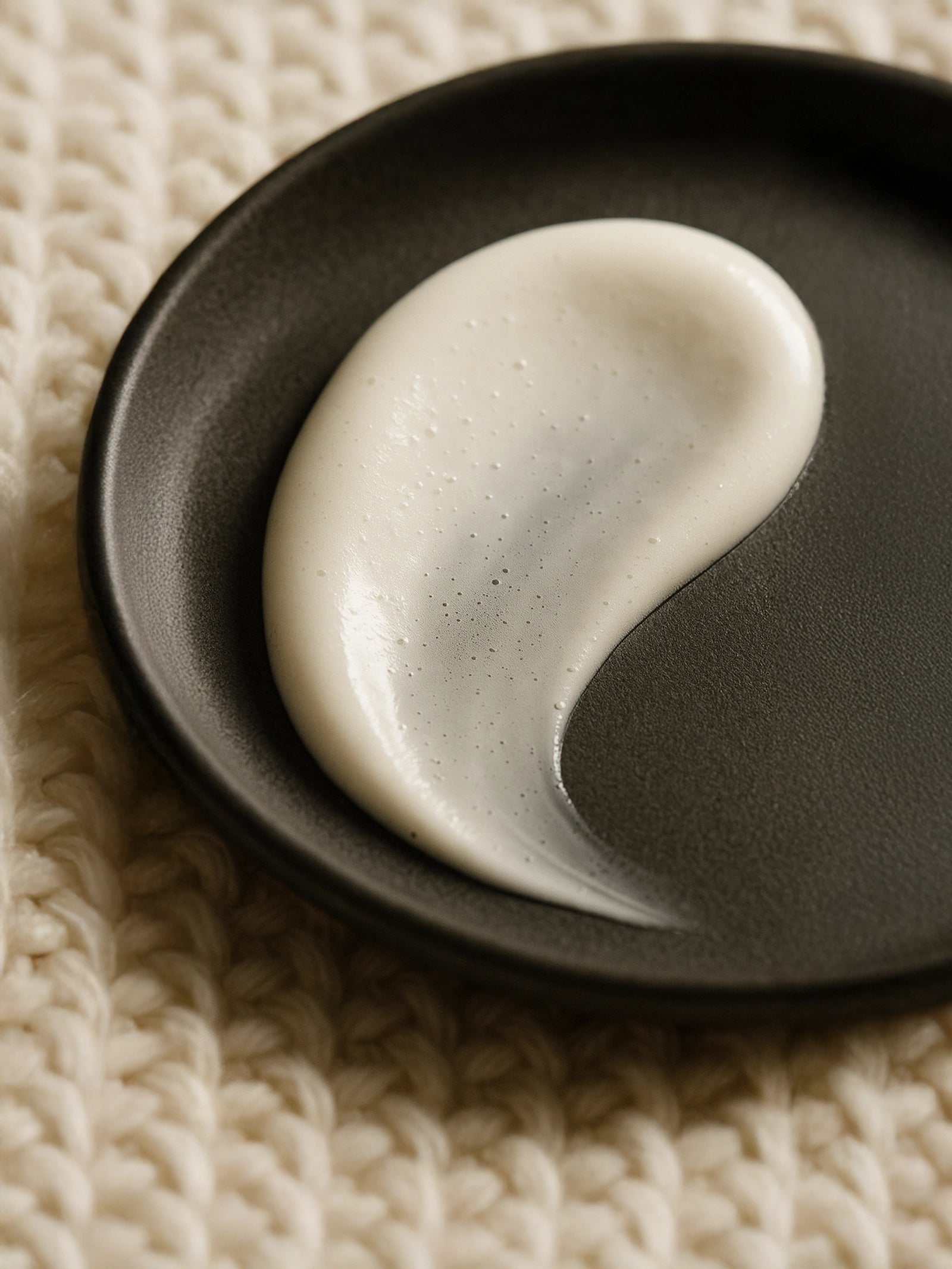 White cream on a black plate on a textured beige surface cleansing milk natural skincare