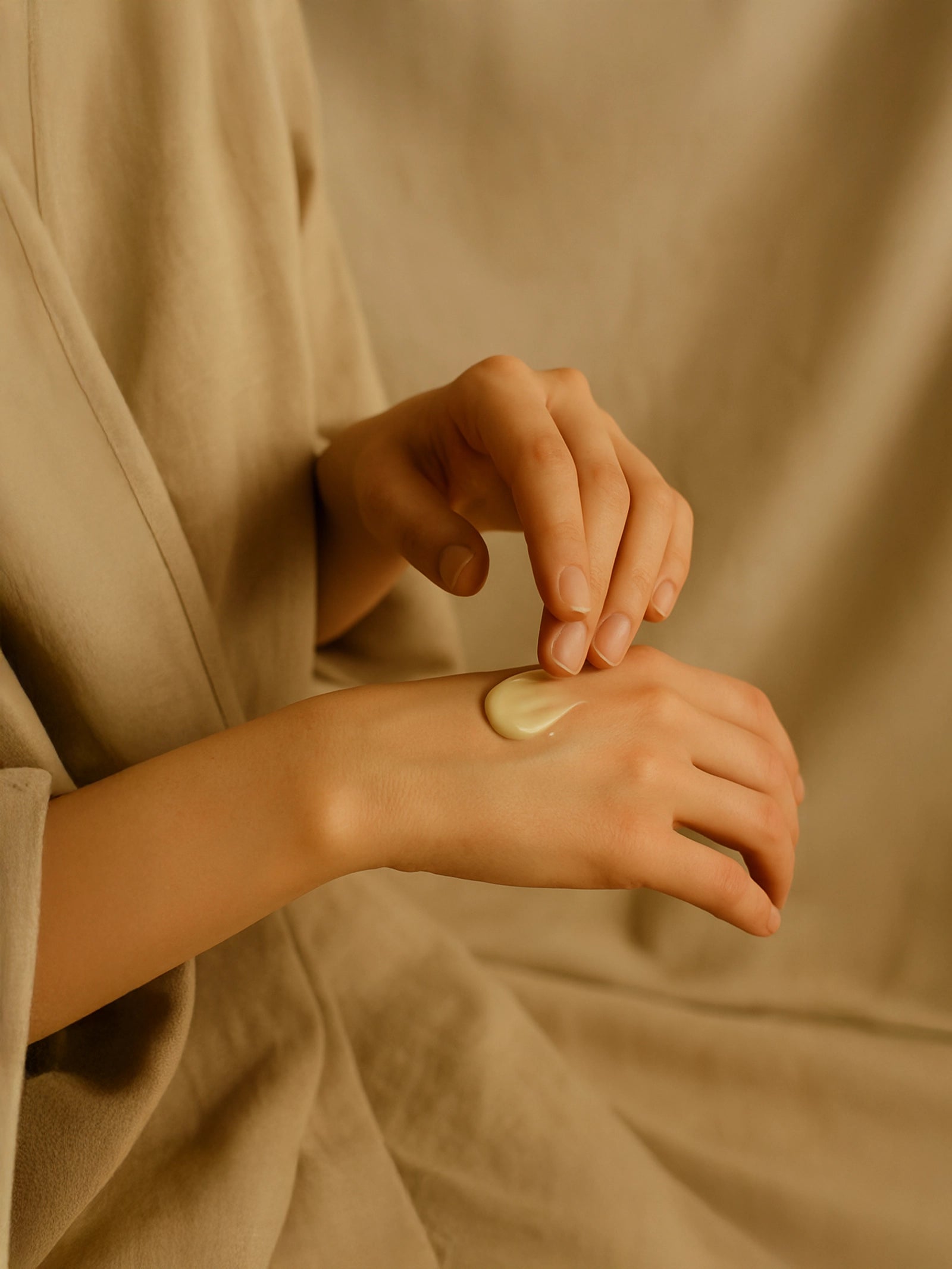Person applying cream to their wrist against a beige background Suvéra Self Care — hands gently spreading a small dollop of cream on the back of the hand, against a soft linen backdrop.
